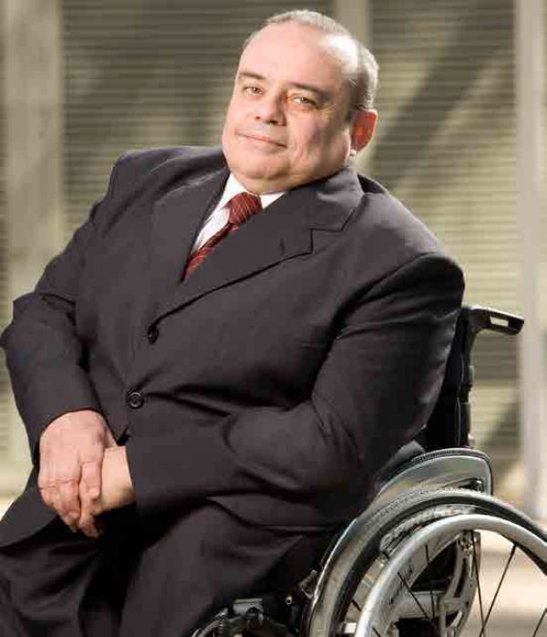 Homem branco. Ele está sentado em uma cadeira de rodas e veste terno cinza com uma camisa branca por baixo e gravata vermelha.